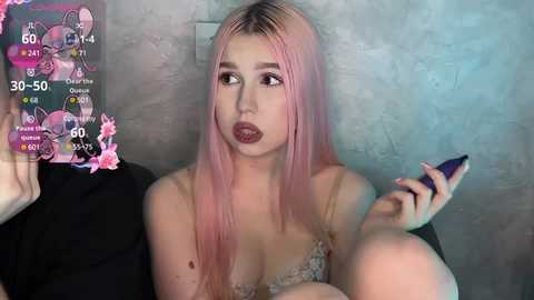 Media: A video of a fair-skinned, blonde-haired woman with pink hair extensions, wearing a lacy bra, looking confused while holding a phone. The background features a textured wall and a black couch.