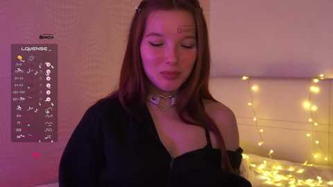 Video of a young woman with fair skin, long brown hair, and a black choker, smiling with closed eyes, standing in a softly lit room with string lights and a bed.
