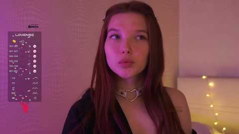 Video of a young Asian woman with long brown hair, wearing a black jacket, standing in a dimly lit room with a \"LOVE\" sign and string lights.