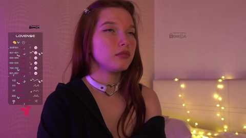 Video of a young woman with long brown hair, wearing a black off-shoulder top and white choker, standing in a dimly lit bedroom with purple and yellow string lights, and a poster of \"Covergirl\" in the background.