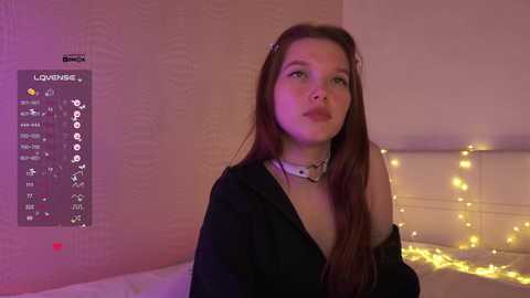 Video of a fair-skinned, red-haired young woman in a black jacket, with a white collar, sitting on a bed adorned with yellow fairy lights. The background features a wall with a \"Love Island\" poster.