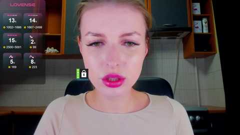 Video of a Caucasian woman with fair skin, light blonde hair, and pink lipstick, sitting in a modern kitchen with wooden cabinets and tiled walls. She looks directly at the camera.