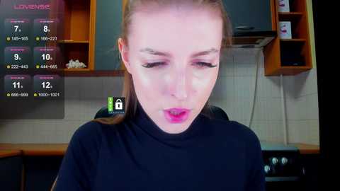 Video of a young, fair-skinned woman with straight, light brown hair tied back, wearing a black turtleneck sweater, in a modern kitchen setting.