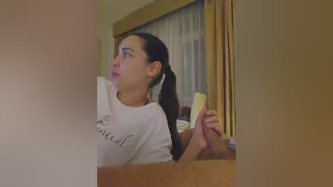 A video of a woman with long dark hair tied back, wearing a white T-shirt with \"Cute\" written on it, holding a banana. She's looking to the side, indoors with yellow curtains and a window in the background.