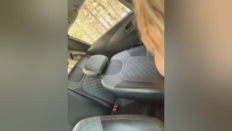 Video of a person in the backseat of a car, with their blonde hair visible, facing the window. The car has black quilted leather seats, and the view outside shows autumn foliage.