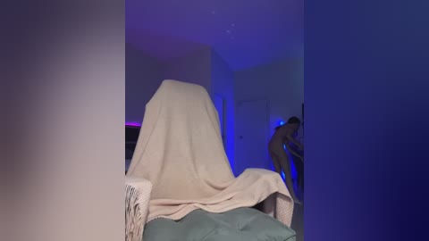Video of a dimly lit bedroom with a beige blanket covering a bed, a person in the background partially visible, blue ambient lighting, and a white wall.
