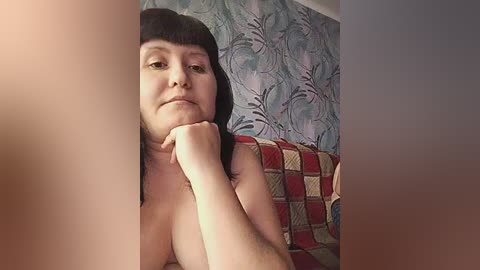 A video of a topless, fair-skinned woman with shoulder-length black hair, resting her chin on her hand, against a patterned blue wall and a red and beige quilted couch.
