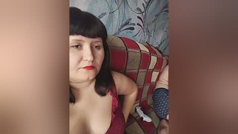 A video of a middle-aged woman with fair skin, black bob haircut, and red lipstick, wearing a low-cut red lace top, sitting on a plaid couch with floral wallpaper background.