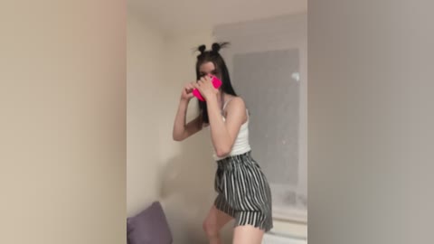 Video of a young woman with fair skin and long black hair in two buns, wearing a white tank top and black-and-white striped shorts, holding a pink phone, indoors with beige walls and a window.