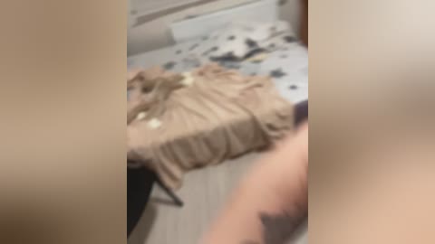 Blurry video of a beige blanket on a bed with a white and black patterned sheet, a partially visible tattooed arm, and a cluttered room background.