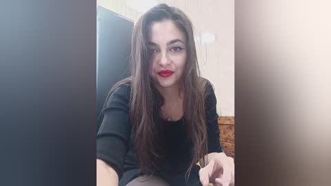 Media: Video of a fair-skinned woman with long, dark hair, wearing red lipstick and a black top, seated in a home setting with a beige wall and wicker chair in the background.