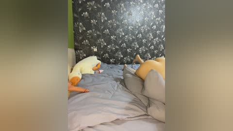 Media: Video of a person lying on a bed with gray sheets, surrounded by plush toys, including a duck and a large bear, against a dark floral-patterned wall.