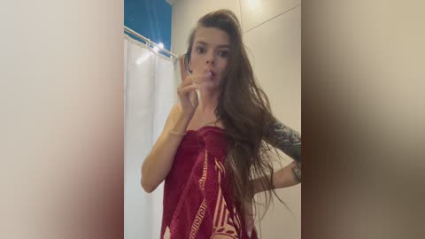 Media: Video of a slender, light-skinned woman with long, wavy brown hair, wearing a red, sheer, strapless dress with gold accents. She has tattoos on her arm, and stands in a bathroom with a blue shower curtain and white tiles.