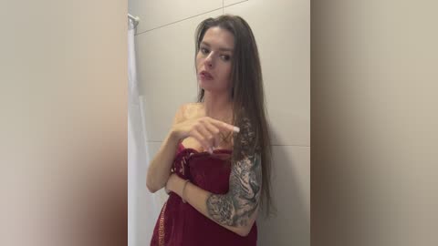 Media: A video of a young woman with long brown hair and fair skin, wearing a red dress, standing in a tiled bathroom. She has a tattoo sleeve on her left arm, and her right arm is raised, pointing.