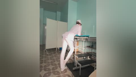 Media: A video of a nurse in a sterile medical room, wearing white scrubs, a hat, and gloves, bending over a patient's bed. The room has light blue walls, a patterned floor, and medical equipment.