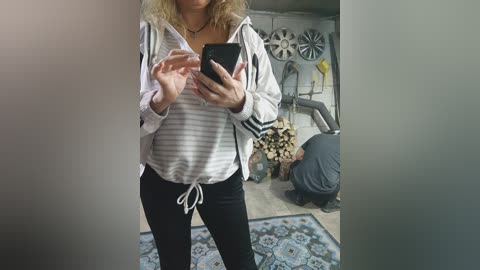 Media: Video of a woman with wavy blonde hair, wearing a striped hoodie and black pants, taking a mirror selfie in a cluttered room with a fan, laundry, and a rug.