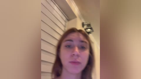 Media: Video of a young woman with fair skin and brown hair, standing in a dimly lit room with beige walls and a white shuttered window. She has a neutral expression and appears to be indoors.