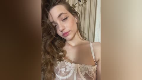 A video of a young woman with long, wavy brown hair, fair skin, and delicate facial features. She wears a sheer, lace bralette, posing indoors with a soft, natural light.