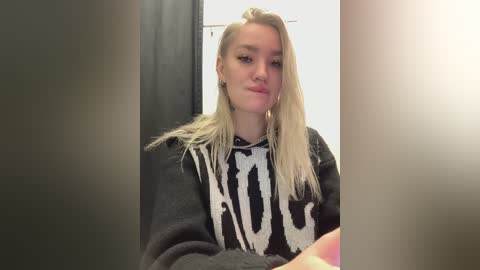 A video of a young woman with long, straight blonde hair, wearing a black sweater with a white abstract pattern, seated indoors.