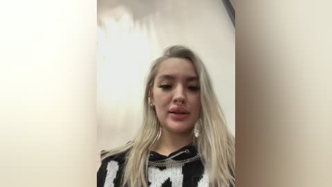 Video of a young woman with long, straight platinum blonde hair, wearing a black and white patterned sweater. She has fair skin and is standing in front of a beige wall, with a slight smile.