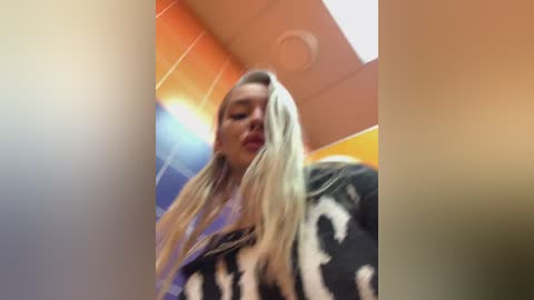 Video of a blonde woman with long hair, wearing a black and white patterned jacket, standing in a colorful, abstract hallway with orange and blue walls.