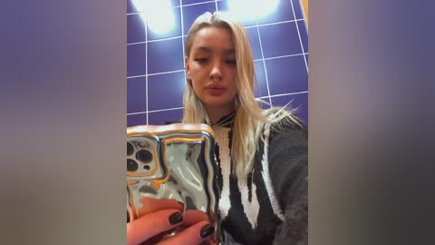 A video of a young woman with long, platinum blonde hair, fair skin, and light makeup, taking a selfie in a bathroom with purple tiled walls and fluorescent lighting.