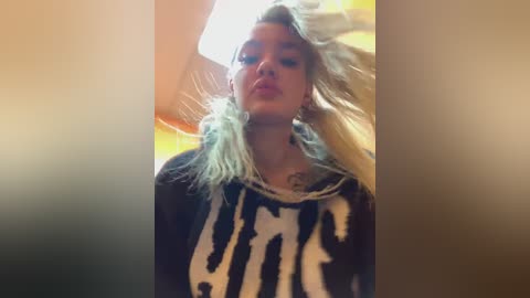 Video of a young woman with long, tousled platinum blonde hair, wearing a black graphic tee. Background is blurred, suggesting an indoor setting with warm lighting.