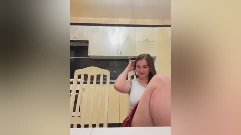 Media: A video of a young, fair-skinned woman with long brown hair, wearing a white top and red shorts, sitting on a wooden chair in a modern kitchen with cream cabinets and black countertops.