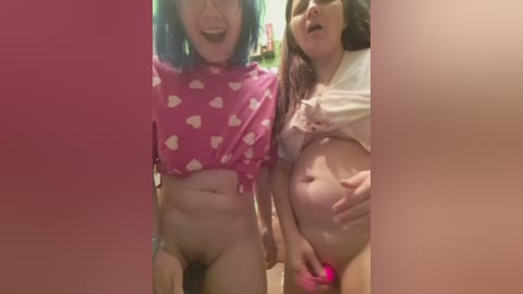 A video of two women, one with blue hair and pink polka-dot crop top, the other with brown hair and white shirt, both nude, in a bathroom.