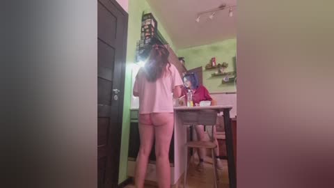 Video of a young woman with long brown hair, wearing a white shirt, standing in a small kitchen, revealing her buttocks.
