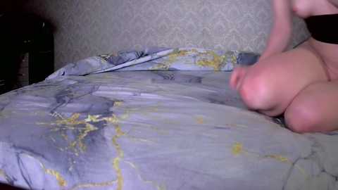 Video of a nude woman with pale skin, kneeling on a bed with a blue and yellow patterned sheet, in a dimly lit room with a patterned wallpaper.