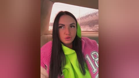 Video of a young woman with long black hair, wearing a bright pink and green jacket, looking surprised or shocked, inside a car with beige seats.