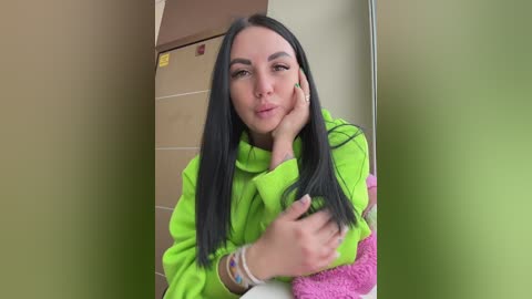 A video of a young woman with long, straight black hair, fair skin, and a slight smile. She's wearing a bright neon green hoodie and is resting her chin on her hand, looking relaxed in a dimly lit room with beige and brown walls.