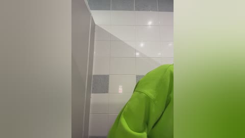 Video of a person wearing a bright green hoodie, partially visible, standing in a bathroom with white tiled walls and a glass shower door.