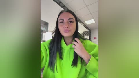 Video of a young woman with long black hair, fair skin, and a slight pout, wearing a bright green hoodie. She stands in a modern office with white walls, fluorescent lights, and a blurred background.