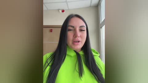 A video shows a young woman with long, straight black hair, wearing a bright neon green hoodie, standing in a dimly lit hallway with beige walls and a ceiling light.