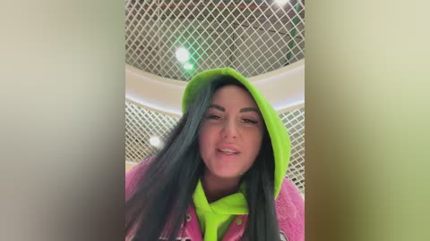 Video of a smiling woman with long black hair, wearing a neon green hoodie and scarf, standing in front of a metal mesh wall with a circular pattern.