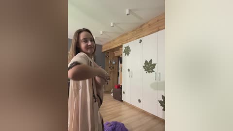 Media: Video of a Caucasian woman with long brown hair, wearing a beige dress, standing in a modern, light-wooden-floored bedroom with white wardrobe doors adorned with green leaf decals.