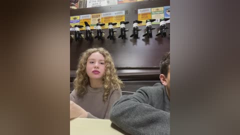 Media: Video of a woman with curly blonde hair and pink lipstick, wearing a beige sweater, seated in a dimly lit restaurant with black-and-white toy police figures mounted on the wall.