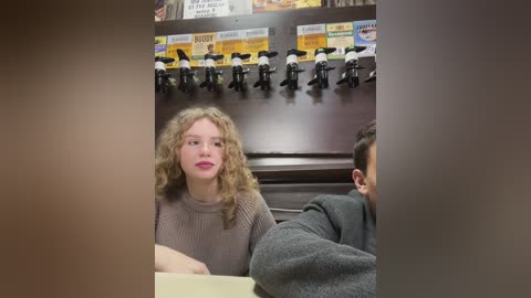 Media: Video of a woman with curly blonde hair, wearing a beige knit sweater, sitting next to a man in a grey sweater in a restaurant. A menu with \"Burger\" and \"Fries\" is visible behind them.