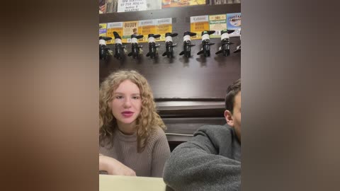 Media: Video of a young woman with curly blonde hair, wearing a brown turtleneck, seated in a booth at a restaurant. Behind her, black bike helmets hang on hooks above a menu board.