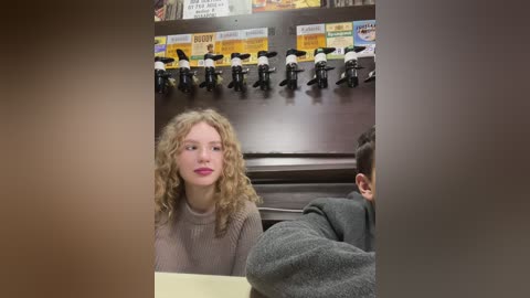 Media: Video of a blonde woman in a beige sweater seated at a table in a dimly lit coffee shop, next to a man with short brown hair, with black coffee machines mounted on the wall.