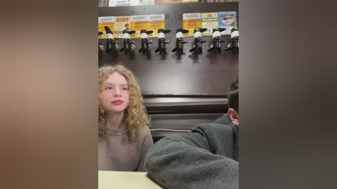 Media: Video of a blonde woman in a beige sweater, sitting in a coffee shop with a toy display of black and white figures in the background.