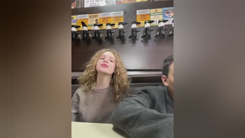Media: A video of a young woman with curly hair and pink lipstick, wearing a beige sweater, sitting at a table in a restaurant with a dark wooden backdrop and multiple toy figures mounted above.