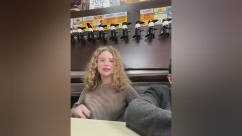 Media: Video of a young Caucasian woman with curly blonde hair, wearing a beige ribbed sweater, seated at a counter with a black and white mug in front of her. Behind her, a dark wooden wall features a menu and toy figurines.