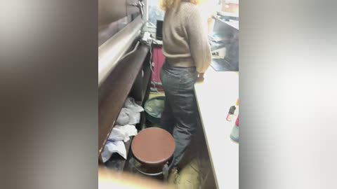 Media: A video shows a woman with blonde hair, wearing a beige sweater and black pants, squatting on a brown stool in a cluttered kitchen with white walls and a tiled floor.