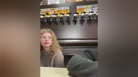 Media: Video of a blonde woman with curly hair, wearing a beige sweater, seated at a counter with a wooden background. A row of black-and-white figurines and colorful signs are mounted above her.