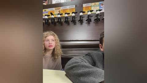 Media: Video of a blonde woman with curly hair, wearing a gray sweater, sitting at a desk in an office with a wooden wall and mounted toy figurines.