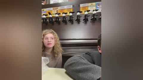 Media: Video of a young girl with curly blonde hair and pink lipstick, wearing a beige sweater, sitting in a booth. Behind her, a row of black mic stands with yellow signs.