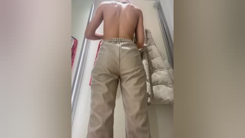 Media: Video of a shirtless person with a muscular back, wearing beige cargo pants, standing in a narrow hallway with a beige jacket hanging on the wall.